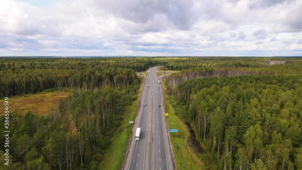 Aerial view of an asphalt road with trucks and cars. The vehicles are going fast, straight ahead. Blue sky with thick white clouds. There is a dense forest with green trees. Warm summer day. UHD. Aerial view of an asphalt road with trucks and cars. The vehicles are going fast, straight ahead. Blue sky with thick white clouds. There is a dense forest with green trees. Warm summer day. UHD.