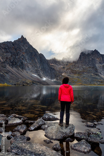 Woman Standing on a Rock at an Alpine Lake surrounded by Rugged Mountains during Fall in Canadian Nature. Taken in Tombstone Territorial Park, Yukon, Canada.