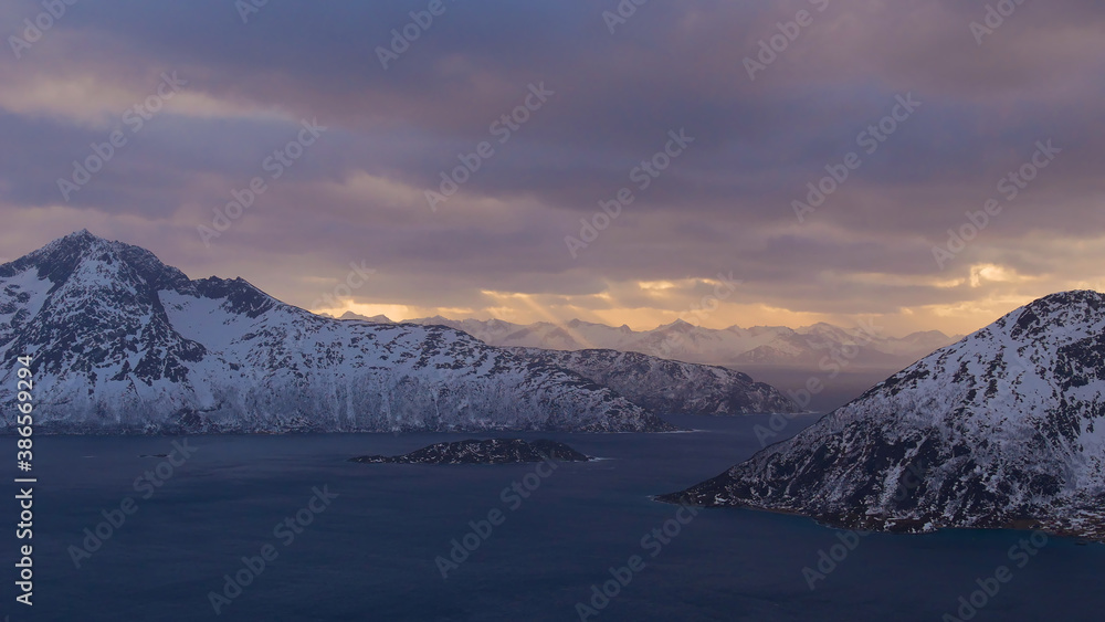 Majestic panorama of afternoon sun shining through clouds with visible sunbeams over the fjords (Sessøyfjord) near Rekvik, Troms og Finnmark, Norway with snow-covered mountains in late winter.