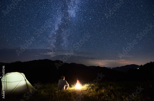 Back view of tourist is relaxing near bonfire and illuminated tent enjoying the silhouettes of mountains under starry sky with bright milky way. Night camping in mountains.
