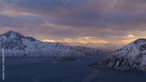 Majestic panorama of afternoon sun shining through clouds with visible sunbeams over the fjords (Sessøyfjord) near Rekvik, Troms og Finnmark, Norway with snow-covered mountains in late winter.