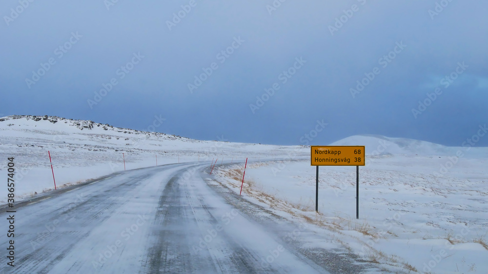 Fototapeta premium Icy road E69 (European route) with road sign displaying the distance to Nordkapp of 68 km near Repvag, Troms og Finnmark, Norway with red snowplow markings and snow-covered landscape in winter time.