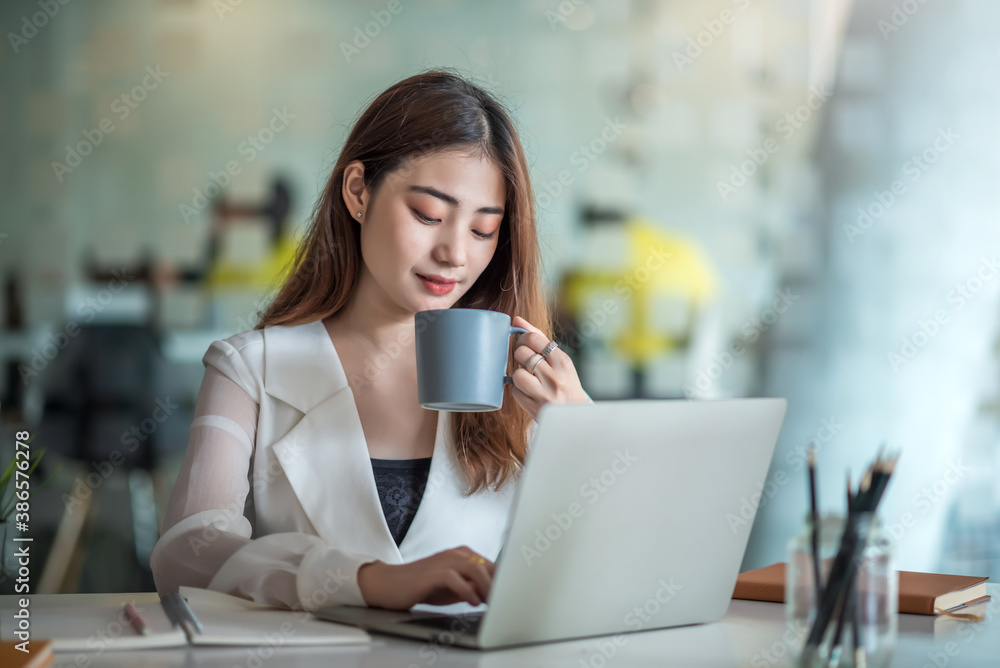 Beautiful asian businesswoman sitting drinking coffee and working happily with a laptop at office