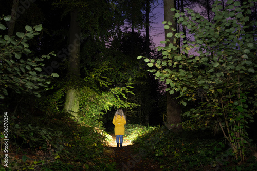 Girl with a yellow coat standing in the dark forrest, searching the path with a flashlight