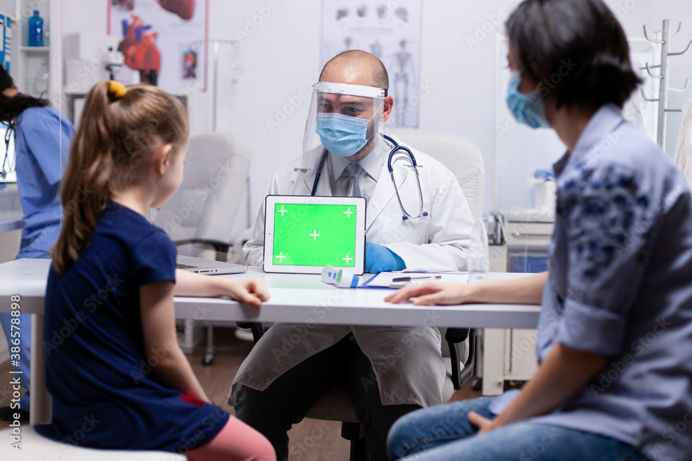 Fototapeta premium Doctor with face mask against coronavirus holding green screen tablet in medical office sitting on the desk. Pediatrician doctor in protection gloves and mask providing clinic medicinal health care