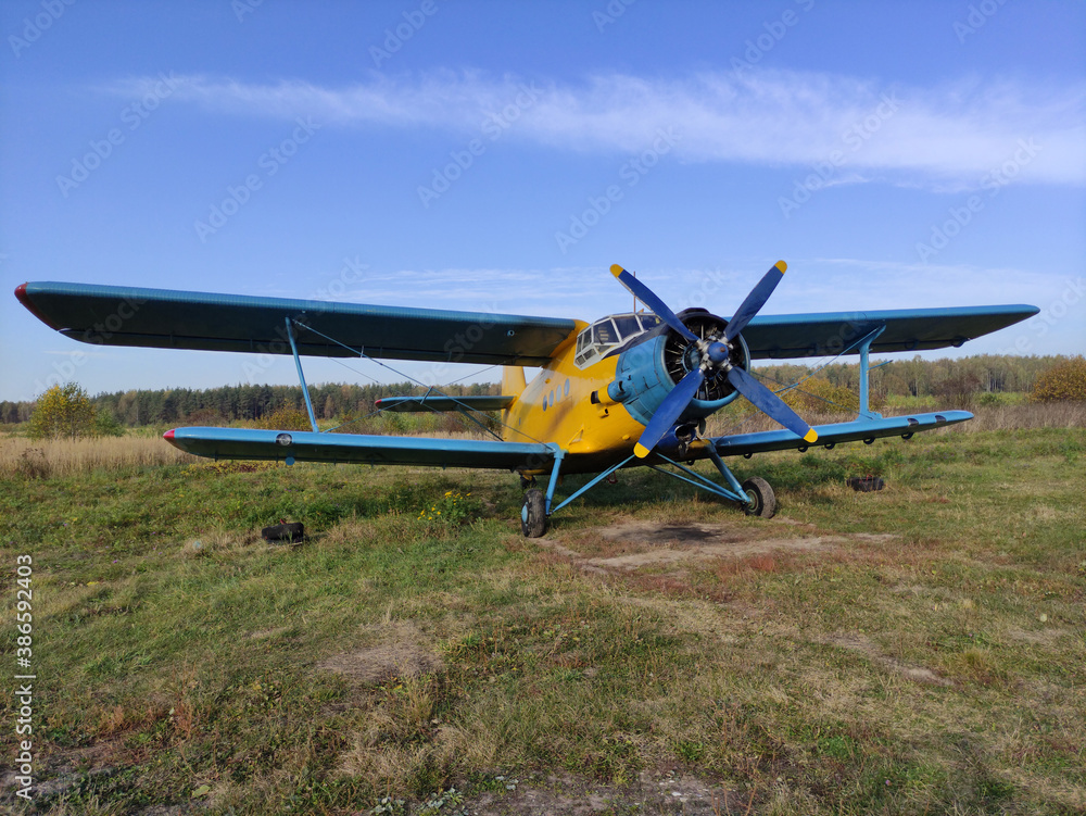 Yellow and blue old biplane plane with a single piston engine and propeller against a blue sky with clouds on the airfield with green grass in summer Yellow and blue old biplane plane with a single piston engine and propeller against a blue sky with clouds on the airfield with green grass in summer