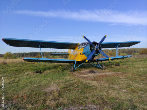 Yellow and blue old biplane plane with a single piston engine and propeller against a blue sky with clouds on the airfield with green grass in summer