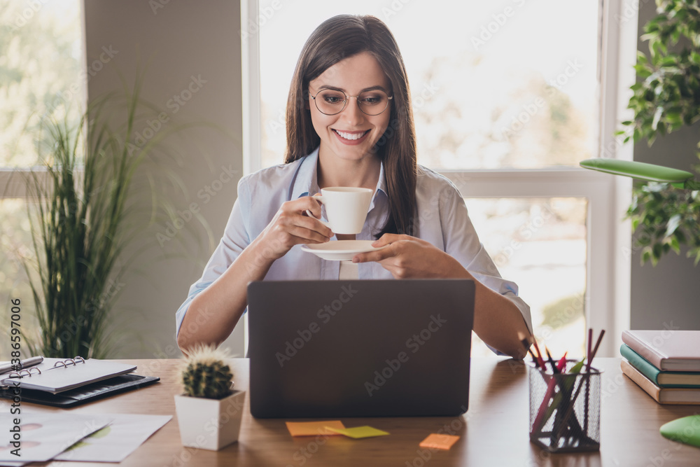 Photo of charming lady sit desktop arms hold cup saucer beaming smile look screen pc wear specs shirt in home office indoors - obrazy, fototapety, plakaty 