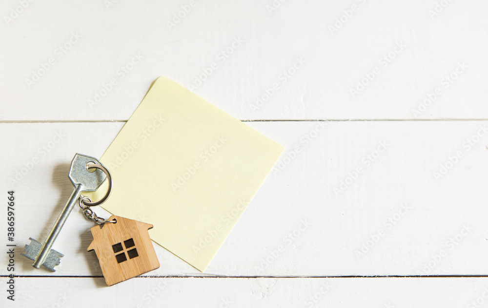 Keychain in the shape of wooden house with key on a white background with a square sheet for notes. Building, design, project, moving to new house, mortgage, rent and purchase real estate. Copy space Keychain in the shape of wooden house with key on a white background with a square sheet for notes. Building, design, project, moving to new house, mortgage, rent and purchase real estate. Copy space