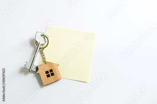 Keychain in the shape of wooden house with key on a white background with a square sheet for notes. Building, design, project, moving to new house, mortgage, rent and purchase real estate. Copy space