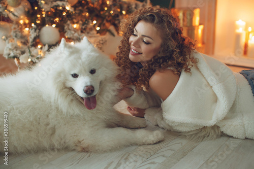 Beautiful woman with a dog hugs, against the background of a fireplace and a Christmas tree with candles. Happy New High quality photo. © nuzza11