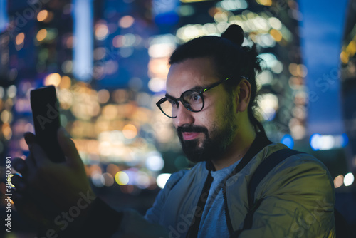 Cute hipster stylish brunette in glasses looks at screen of the smartphone mobile phone in a big city on background of night lights metropolis