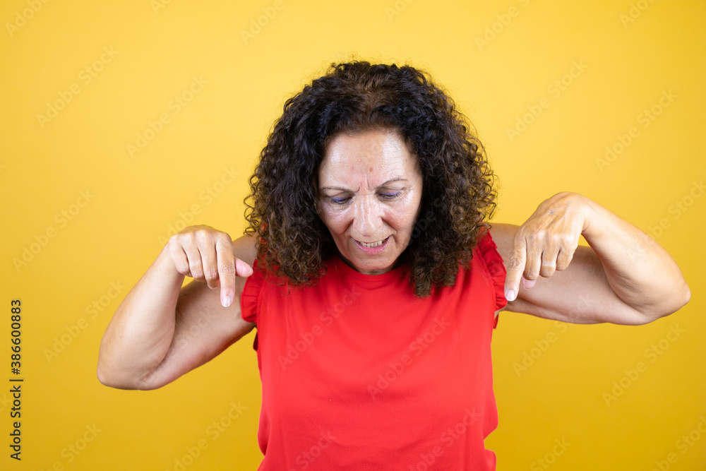 Middle age woman wearing casual shirt standing over isolated yellow background surprised, looking at the camera and pointing down with fingers and raised arms Middle age woman wearing casual shirt standing over isolated yellow background surprised, looking at the camera and pointing down with fingers and raised arms