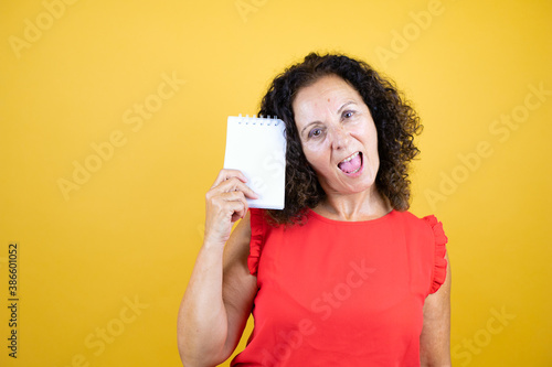 Middle age woman wearing casual shirt standing over isolated yellow background smiling and showing blank notebook in her hand
