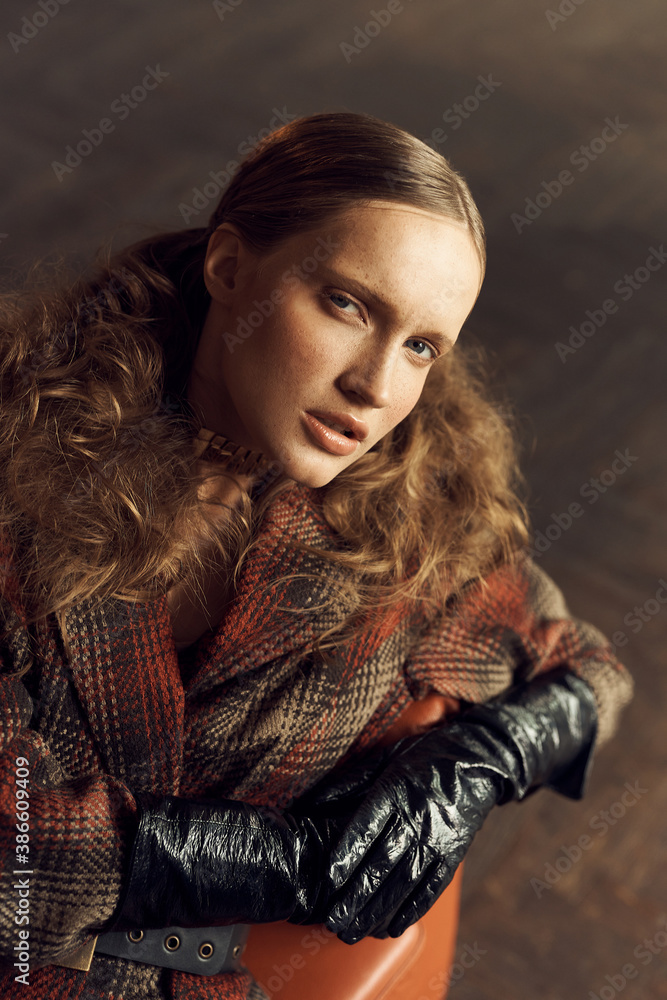 portrait photo of a girl with freckles, brown curly tails in a vintage checkered jacket, she mysteriously looks into the camera sitting on a leather chair