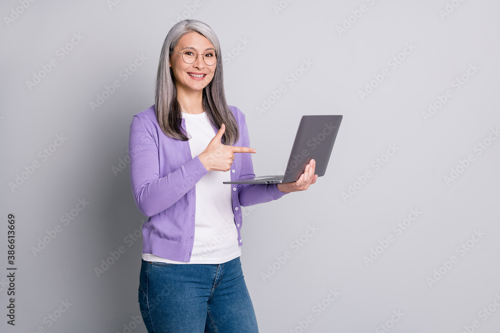 Photo portrait of friendly satisfied smiling old woman holding pointing on laptop with finger isolated on grey color background Photo portrait of friendly satisfied smiling old woman holding pointing on laptop with finger isolated on grey color background