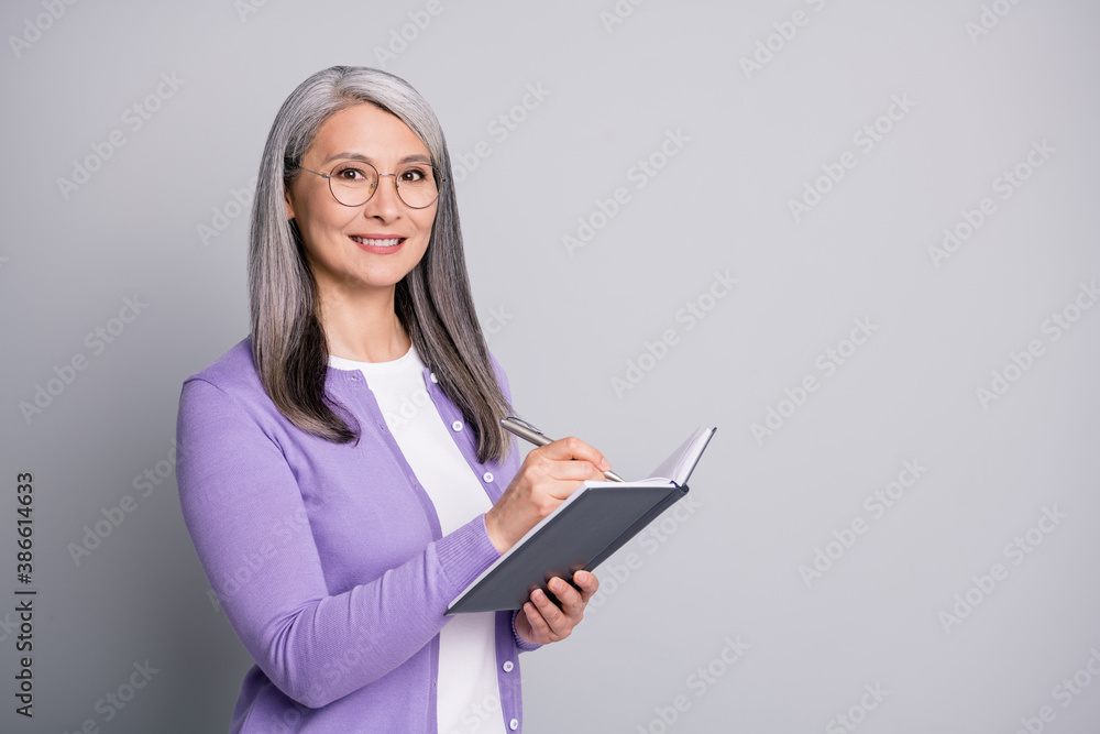 Portrait photo of elder smiling businesswoman writing down notes plans in organizer wearing eyeglasses purple cardigan isolated on grey color background Portrait photo of elder smiling businesswoman writing down notes plans in organizer wearing eyeglasses purple cardigan isolated on grey color background