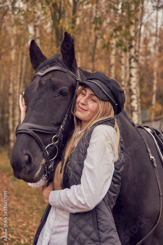 A slender girl holds a horse by the bridle against the background of an autumn forest.