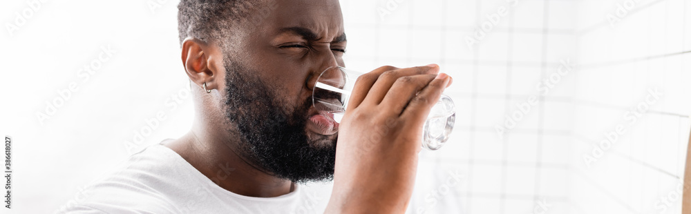 panoramic shot of afro-american man grimacing and drinking water - obrazy, fototapety, plakaty 