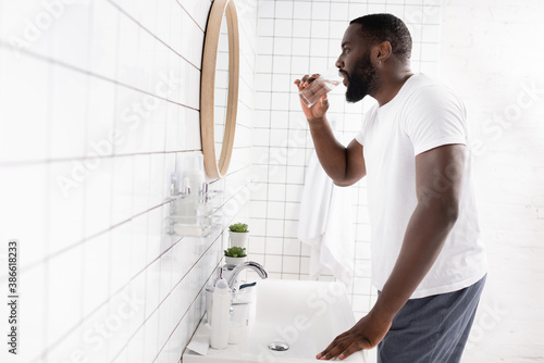 Fotografija side view of afro-american man drinking water and looking in mirror