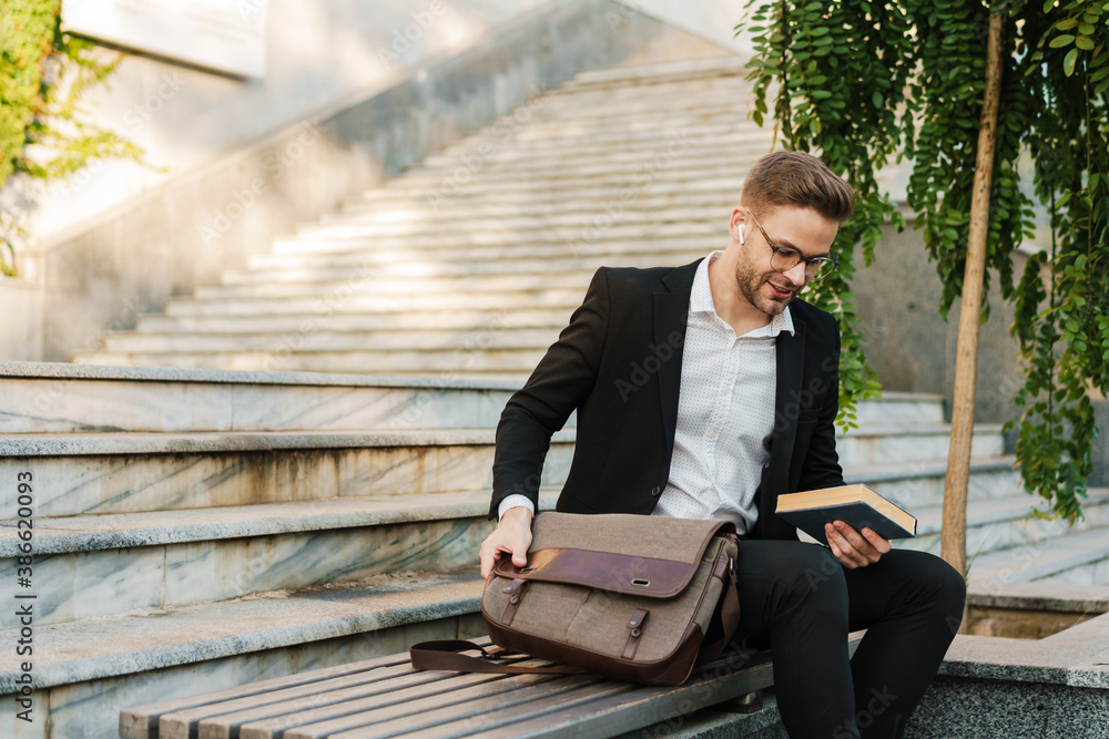 Pleased businessman in earphones holding book while sitting on bench Pleased businessman in earphones holding book while sitting on bench
