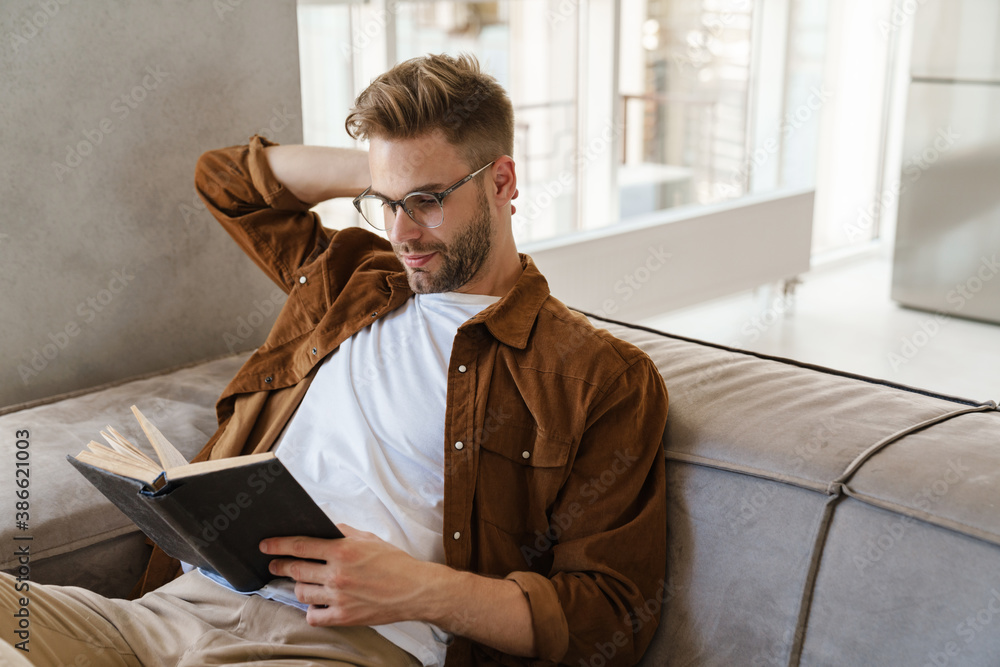 Naklejka premium Handsome focused guy in eyeglasses reading book while sitting on sofa