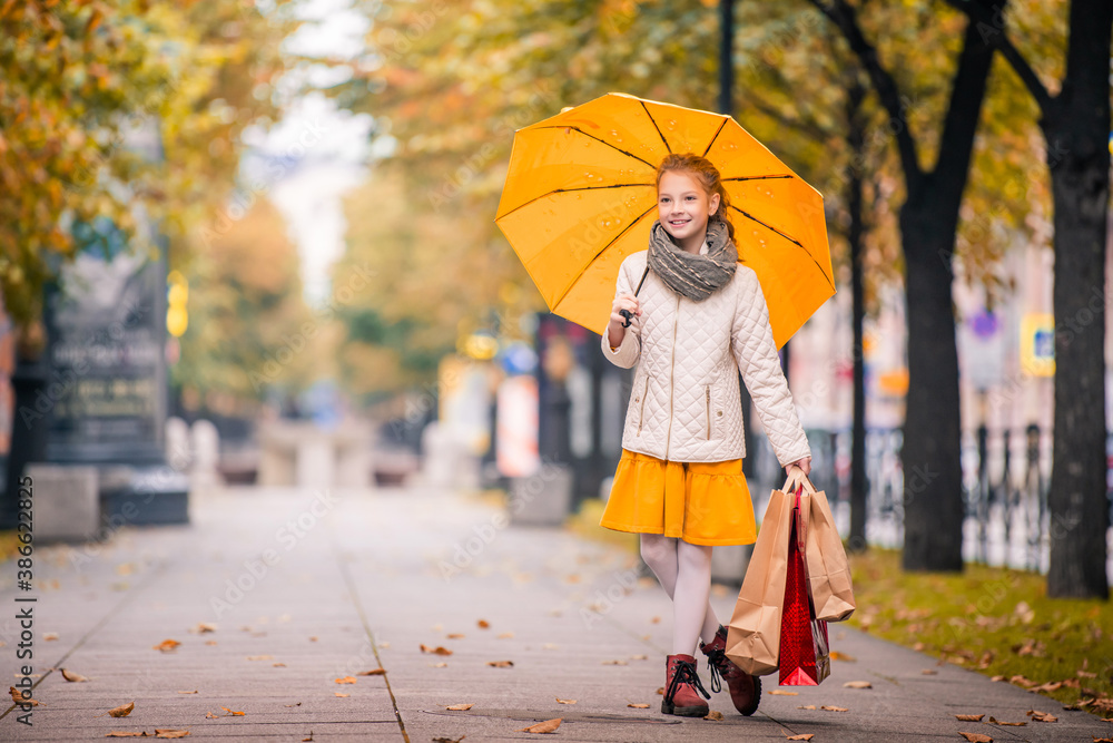 Obraz premium girl with a bright jelly umbrella and with shopping guide walks along the autumn city boulevard smiling happily.