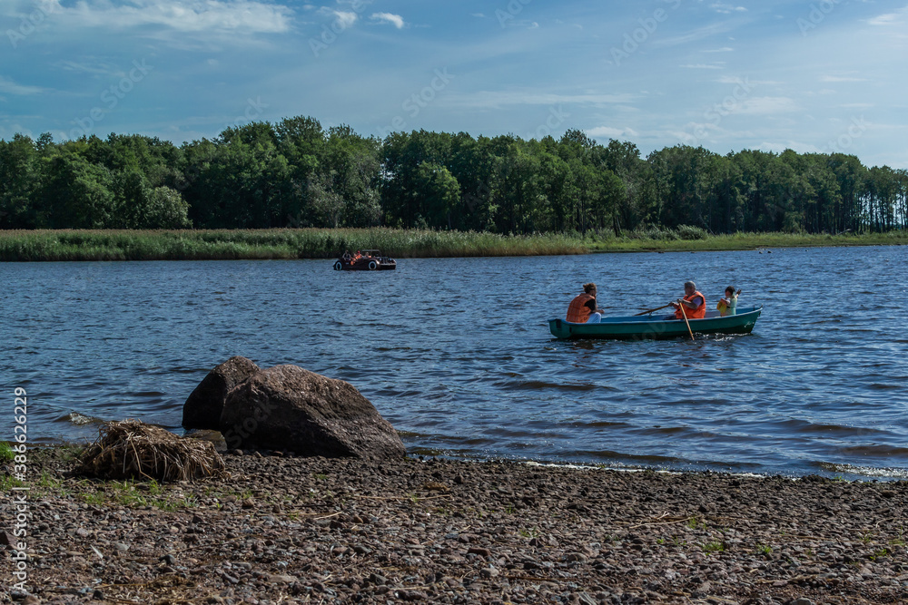 Two small passenger rowing paddle boats with people floats on bay of Baltic sea among green trees, forests in sunny weather. Stones coast. Russia summer Two small passenger rowing paddle boats with people floats on bay of Baltic sea among green trees, forests in sunny weather. Stones coast. Russia summer