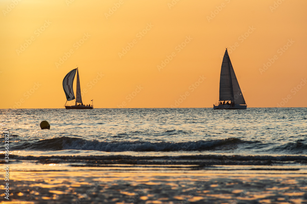 A goldish to orange sky overhangs two sailing boats on a blue greyish sea.