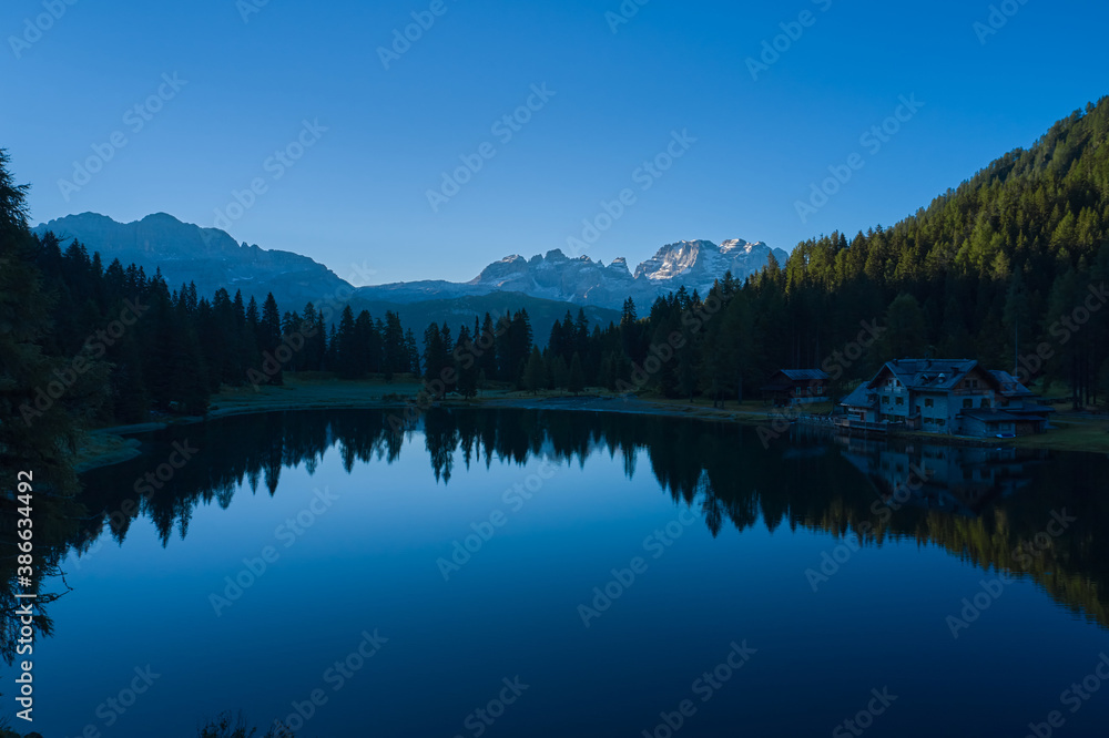 Early morning in the Alps. House in the mountains by a mountain lake. Reflections of trees, mountains, sky in a mountain lake.