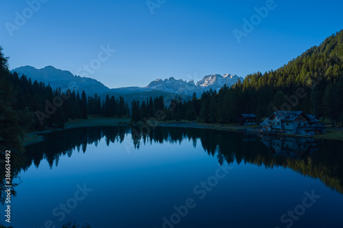 Early morning in the Alps. House in the mountains by a mountain lake. Reflections of trees  mountains  sky in a mountain lake.