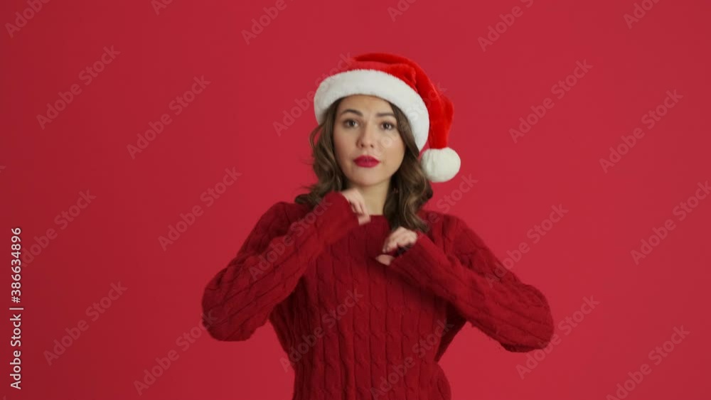 A charming woman wearing a warm red sweater and christmas hat is showing heart and pointing to the viewer standing isolated over red background A charming woman wearing a warm red sweater and christmas hat is showing heart and pointing to the viewer standing isolated over red background