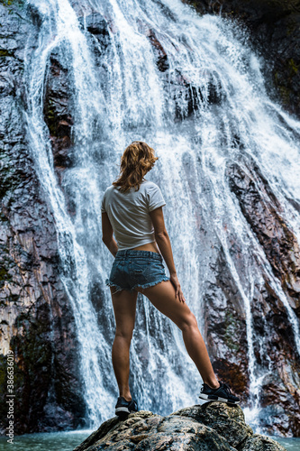 Young caucasian slender woman tourist in casual clothes and sneakers stands on a rock and look at waterfall 