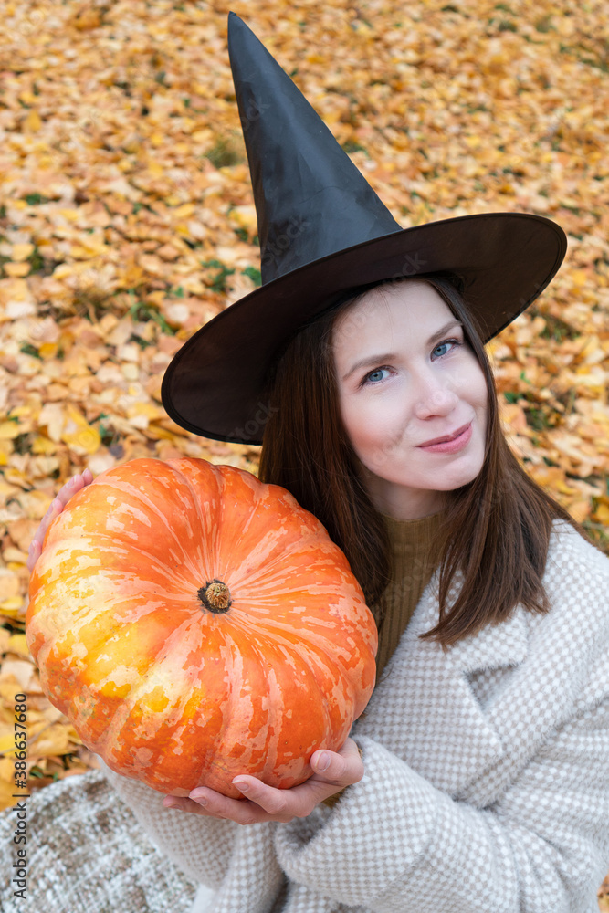 A young beautiful girl in a witch hat and a stylish coat holds a large pumpkin in her hands against a background of yellow foliage. Vertical photo A young beautiful girl in a witch hat and a stylish coat holds a large pumpkin in her hands against a background of yellow foliage. Vertical photo