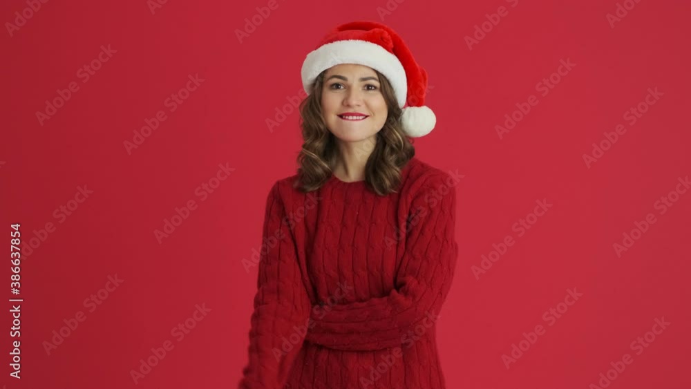 A confident woman wearing a warm red sweater and christmas hat is pointing to the points in the free space standing isolated over red background A confident woman wearing a warm red sweater and christmas hat is pointing to the points in the free space standing isolated over red background