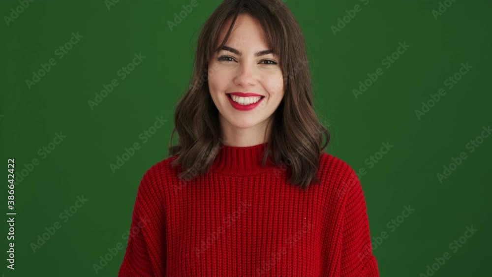 A smiling woman in a red sweater is waving her hand doing hello gesture standing isolated over green background A smiling woman in a red sweater is waving her hand doing hello gesture standing isolated over green background