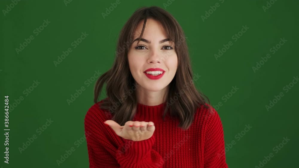 An attractive woman in a red sweater is sending an air kiss to the camera standing isolated over green background An attractive woman in a red sweater is sending an air kiss to the camera standing isolated over green background