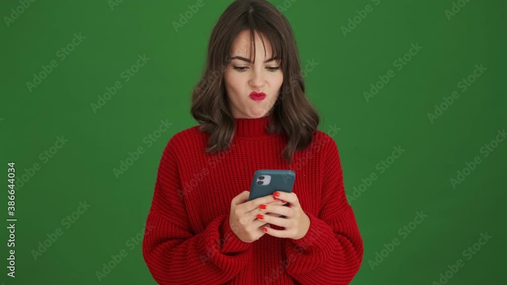 A displeased young woman in a red sweater is using her smartphone standing isolated over green background A displeased young woman in a red sweater is using her smartphone standing isolated over green background