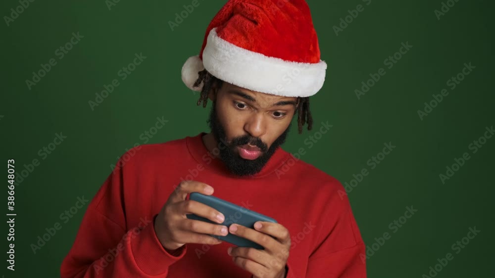 Young excited african man in christmas santa hat playing games by mobile phone isolated over green wall background Young excited african man in christmas santa hat playing games by mobile phone isolated over green wall background