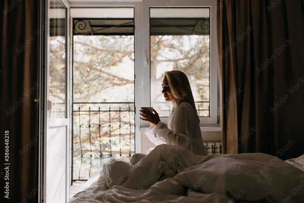 Young woman with a cup of coffee, wearing a pajama, sitting on bed by the window. Young woman with a cup of coffee, wearing a pajama, sitting on bed by the window.