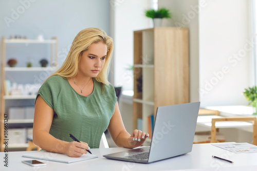 Woman sitting at desk, working online with laptop and making notes