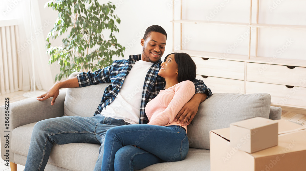 Happy black couple hugging on comfy couch in their new house after unpacking boxes with possessions, copy space