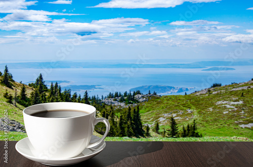 Cup of coffee on the table against mountains background. View of blue sea and green valley in the mountains.