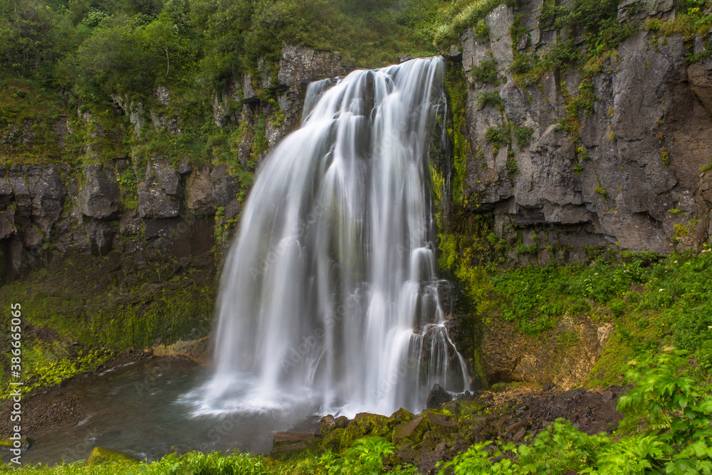 Obraz premium Panoramic view of a small waterfall from a cliff overgrown with trees and bushes