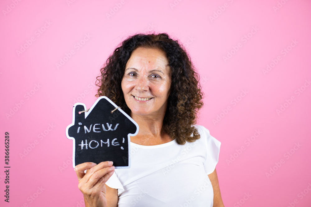 Middle age woman wearing casual white shirt standing over isolated pink background smiling and holding house blackboard with new home word message Middle age woman wearing casual white shirt standing over isolated pink background smiling and holding house blackboard with new home word message
