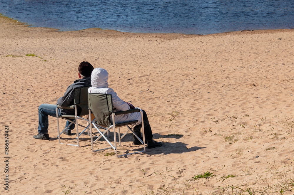 The couple sits on folding chairs on the beach on a sunny cold day.