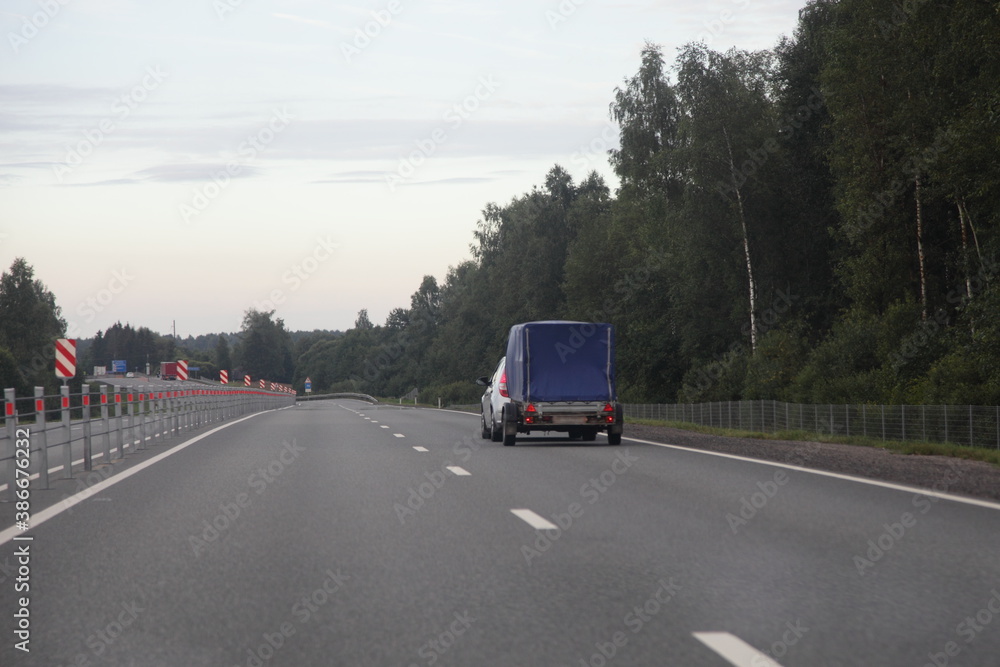 Car with blue tented single axle trailer drive on dry asphalted suburban empty highway road on forest background an summer evening, back view Car with blue tented single axle trailer drive on dry asphalted suburban empty highway road on forest background an summer evening, back view