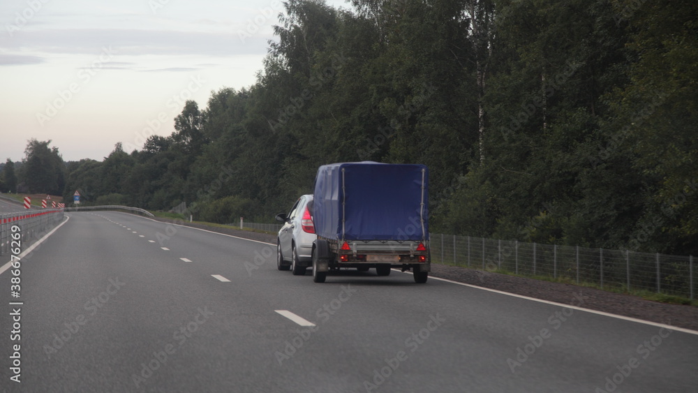 Compact passenger car with awning single axle trailer drive on dry asphalted empty suburban highway road on forest background an summer evening, backside view Compact passenger car with awning single axle trailer drive on dry asphalted empty suburban highway road on forest background an summer evening, backside view
