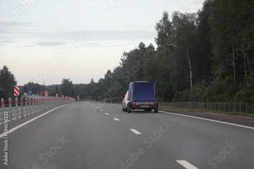 Car with blue tented single axle trailer drive on dry asphalted suburban empty highway road on forest background an summer evening, back view