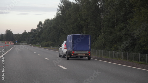 Compact passenger car with awning single axle trailer drive on dry asphalted empty suburban highway road on forest background an summer evening, backside view